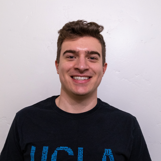 Young man in a black UCLA t shirt standing against a white wall and smiling while facing the camera.